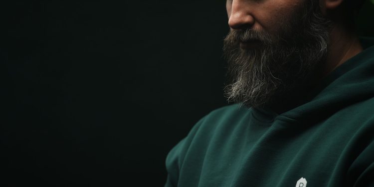 Thoughtful bearded man wearing a dark green hoodie in a moody, low-light setting.