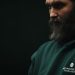 Thoughtful bearded man wearing a dark green hoodie in a moody, low-light setting.