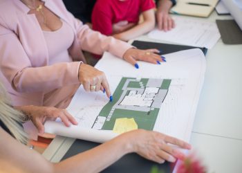 A real estate agent in a pink blazer shows floor plans to clients, discussing property details.