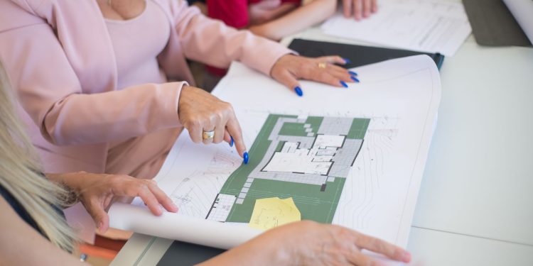 A real estate agent in a pink blazer shows floor plans to clients, discussing property details.