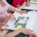A real estate agent in a pink blazer shows floor plans to clients, discussing property details.