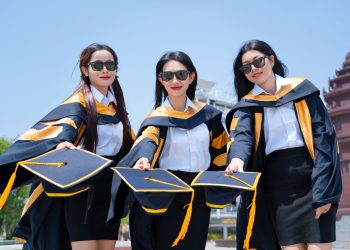Three female graduates in gowns holding caps in celebration outdoors on a sunny day.