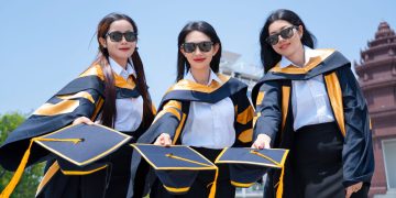 Three female graduates in gowns holding caps in celebration outdoors on a sunny day.