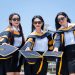 Three female graduates in gowns holding caps in celebration outdoors on a sunny day.