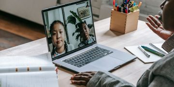 From above of crop confident African American female teacher sitting in classroom and teaching multiethnic pupils remotely on laptop during coronavirus pandemic
