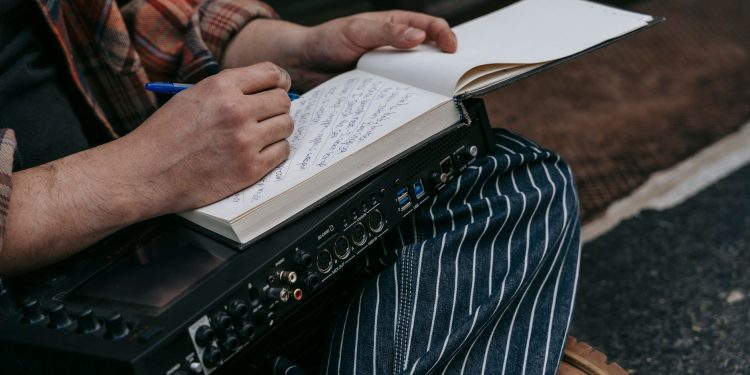 Close-up of a person sitting with a notebook, writing music notes, and holding audio equipment.