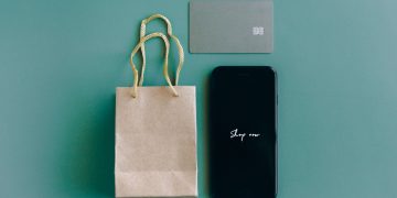 Top view of smartphone with 'Shop now' text, credit card, and paper bag on green background.