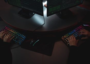 Two people typing on RGB keyboards with code on screens, indicating a cybersecurity environment.