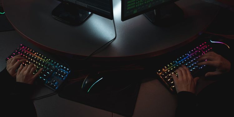 Two people typing on RGB keyboards with code on screens, indicating a cybersecurity environment.