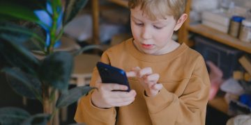 A young boy in a brown sweater uses a smartphone near a potted plant indoors.