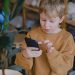 A young boy in a brown sweater uses a smartphone near a potted plant indoors.