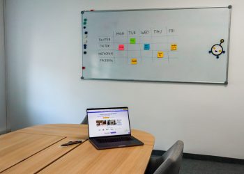 Office setup featuring a conference table, laptop, and organized whiteboard schedule, highlighting a tech-driven workspace.
