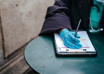 Crop anonymous ethnic female physician in disposable glove with paper on clipboard working at table in daylight