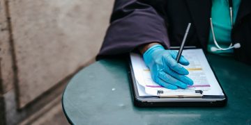 Crop anonymous ethnic female physician in disposable glove with paper on clipboard working at table in daylight