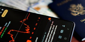 Close-up of a smartphone displaying a stock market app alongside a US passport and currency.