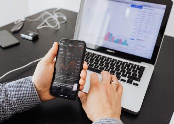 Person analyzes stock data with a smartphone and laptop, indoors.