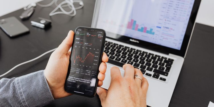 Person analyzes stock data with a smartphone and laptop, indoors.