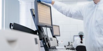 Researcher in PPE reviews data on a digital screen in a modern laboratory setting.
