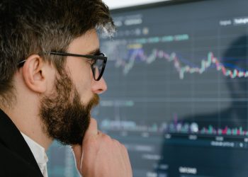 A focused man with glasses studies stock market graphs on a screen, pondering insights.