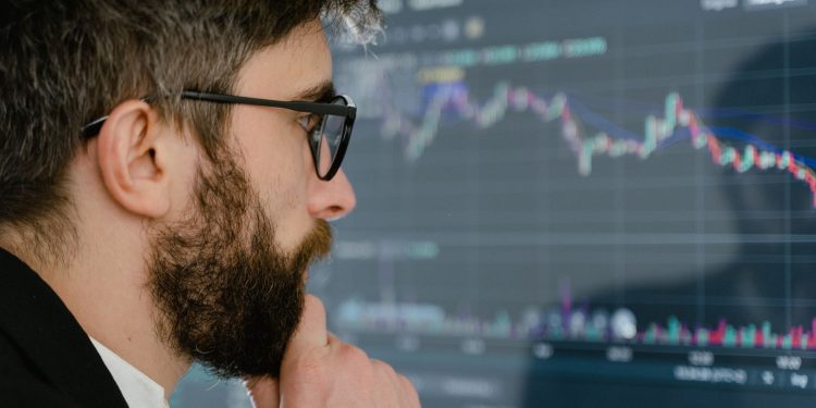 A focused man with glasses studies stock market graphs on a screen, pondering insights.