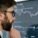 A focused man with glasses studies stock market graphs on a screen, pondering insights.