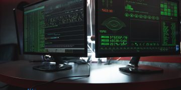 Close-up of dual computer monitors with green coding interfaces in a dark room, highlighting cyber security themes.