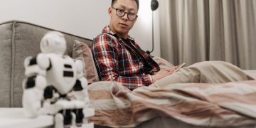 Asian man in plaid shirt sitting on bed, observing a robot toy in a cozy bedroom setting.
